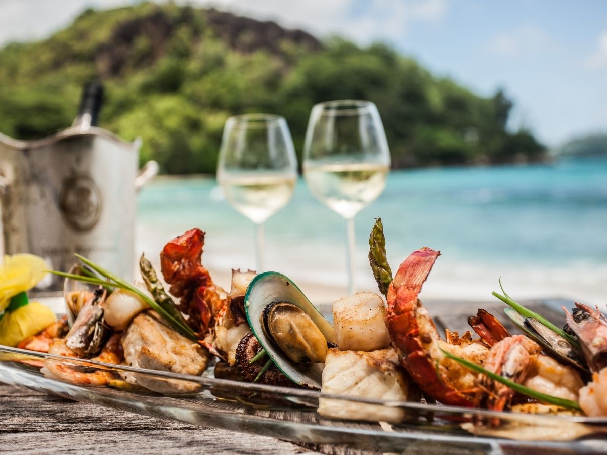 Platter of fresh seafood and two glasses of white wine set on a table by the ocean.